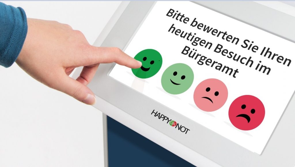 Feedback-Stand mit Tablet für die Bewertung einer Behörde. Ein Finger zeigt bei der Umfrage "Bitte bewerten Sie Ihren heutigen Besuch im Bürgeramt" auf ein sehr zufriedenes Smiley.