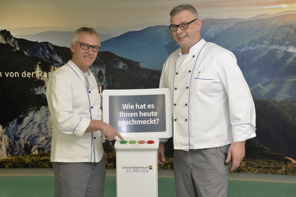 In der Klinik verbessert das Küchenteam das Essen, indem ein Smiley Terminal die Zufriedenheit erfasst und auswertet. Zwei Mitarbeiter drücken auf den Knopf des Terminals.