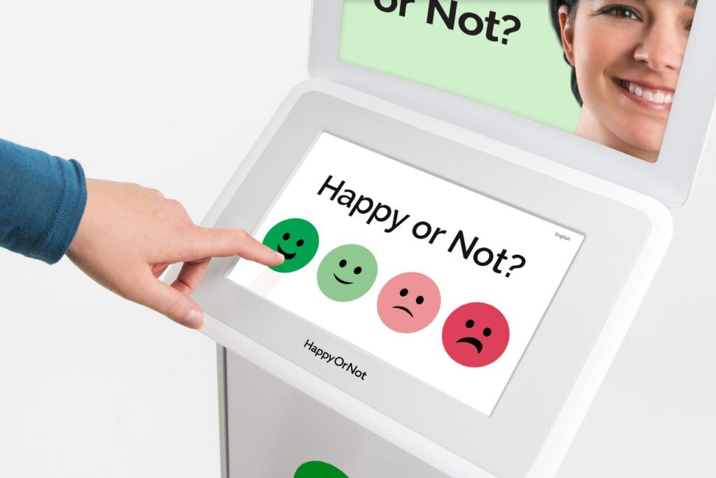 Smiley Touch Gerät mit Frageschild und der vierstufigen Smiley Bewertungsskala mit einer linken Hand, die auf das sehr fröhliche grüne Gesicht drückt.