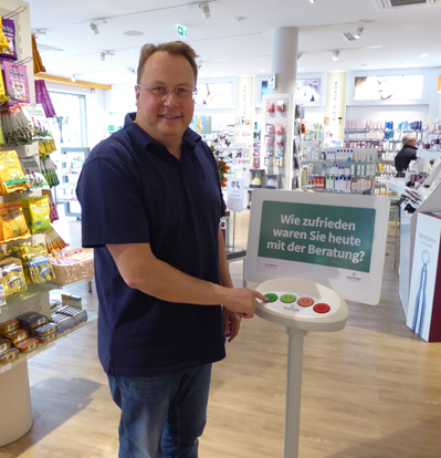Feedbackübermittlung per Knopfdruck in einer von vier Apotheken. Der Eigentümer drückt auf das sehr glückliche Smiley-Gesicht.