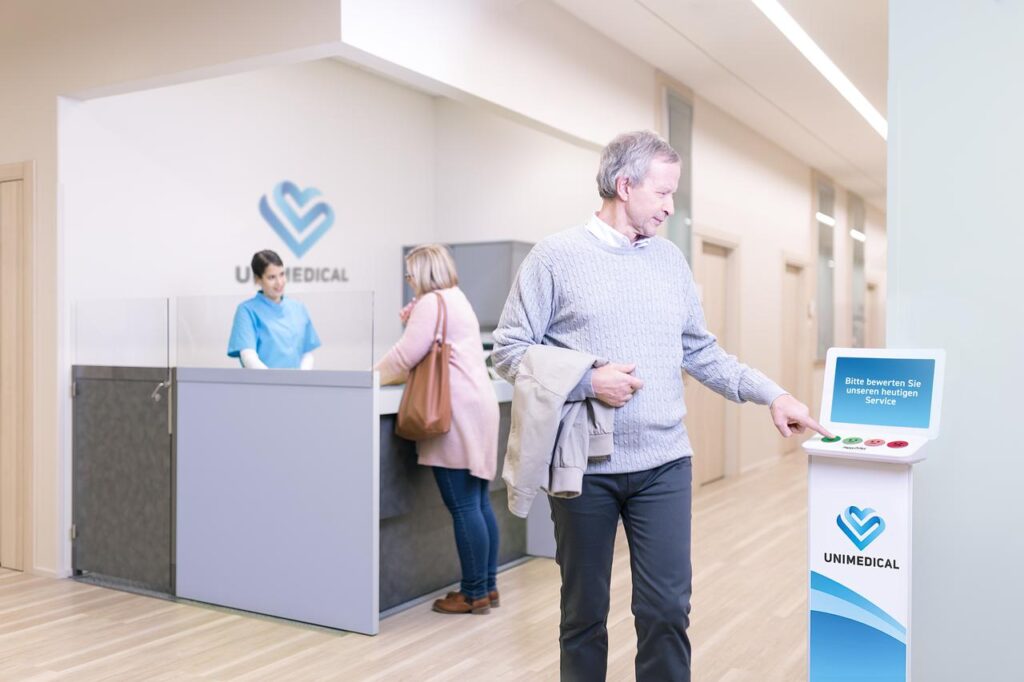Ein Patient bewertet das Aufnahmemanagement einer Gesundheitseinrichtung durch Betätigen eines Tastendrucks einer Bewertungsstele. Er wählt das sehr zufriedene Smiley.