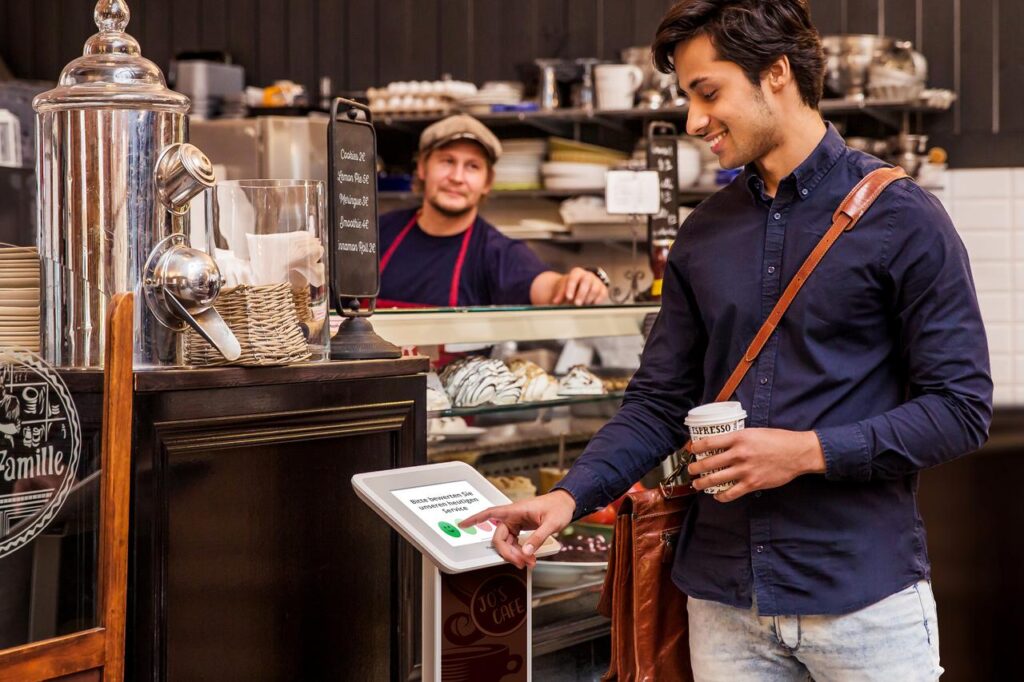 Lächelnder Mann in einer Bäckerei, der einen Kaffee in der Hand hält und das HappyOrNot Smiley Touch Terminal bedient.