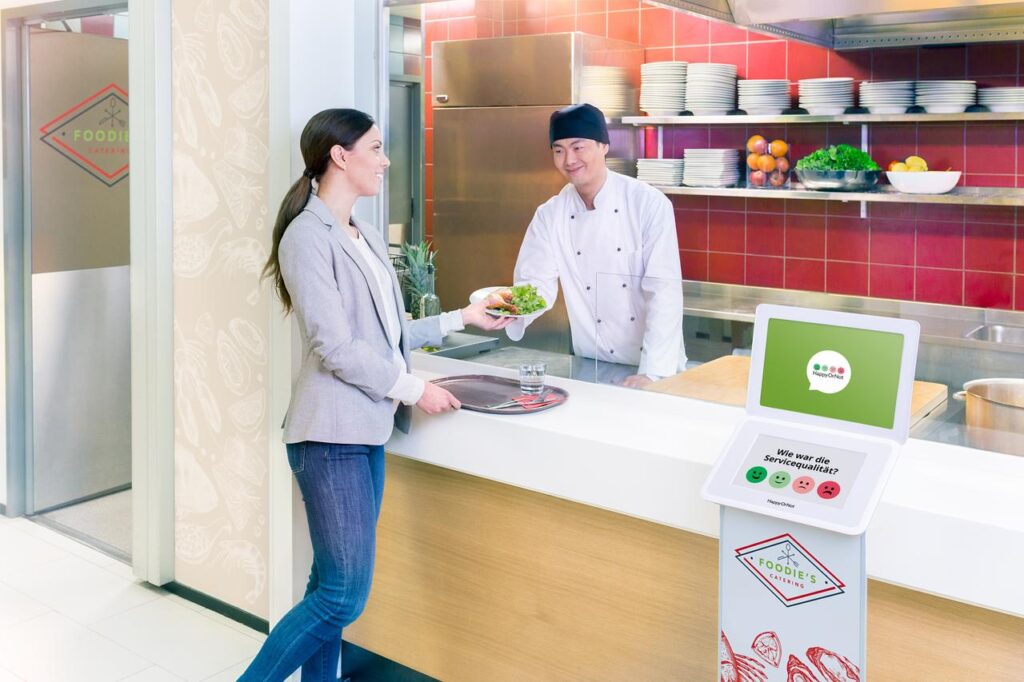 Ein Gast nimmt einen Teller mit Essen, den sie vom Koch einer Kantine erhält. Im Vordergrund ist ein Tablet mit einer digitalen Befragung aufgestellt.